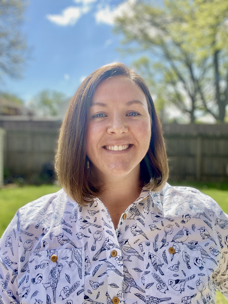 Photo of Tracey Berg Fulton, a white woman with shoulder length brown hair. She is wearing a white shirt with blue birds printed on it and standing outside on a sunny day smiling at the camera.